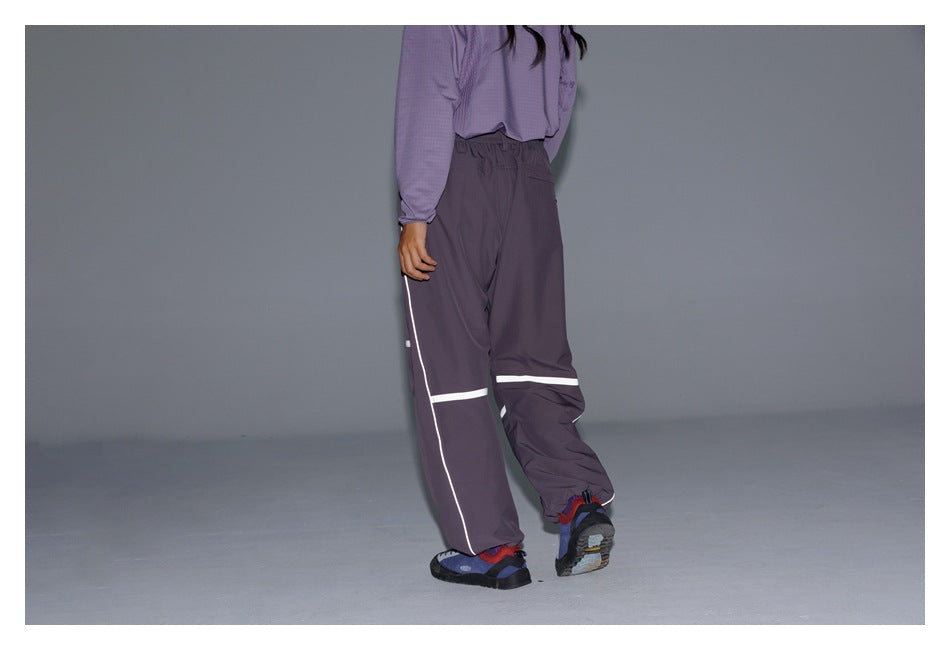 Three-quarter rear view of a model wearing dark plum loose pants with white reflective bands and side piping.