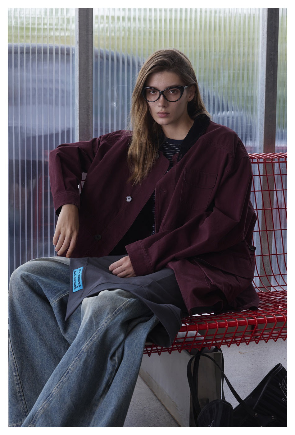 Seated model wearing an oversized burgundy jacket with a black collar and flap pockets.