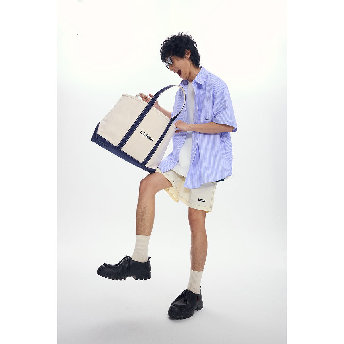 Model in a blue striped short-sleeve shirt and beige shorts carries a cream tote with navy trim.