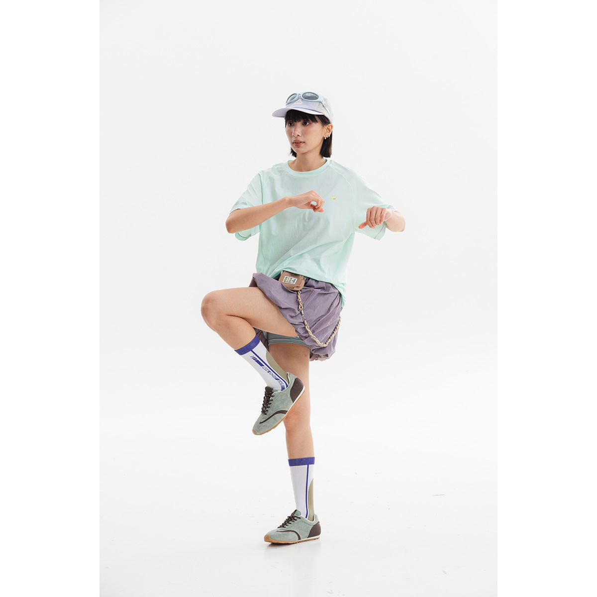 Model wearing a loose pale mint short-sleeve T-shirt with a small chest emblem, purple shorts, and a white cap.