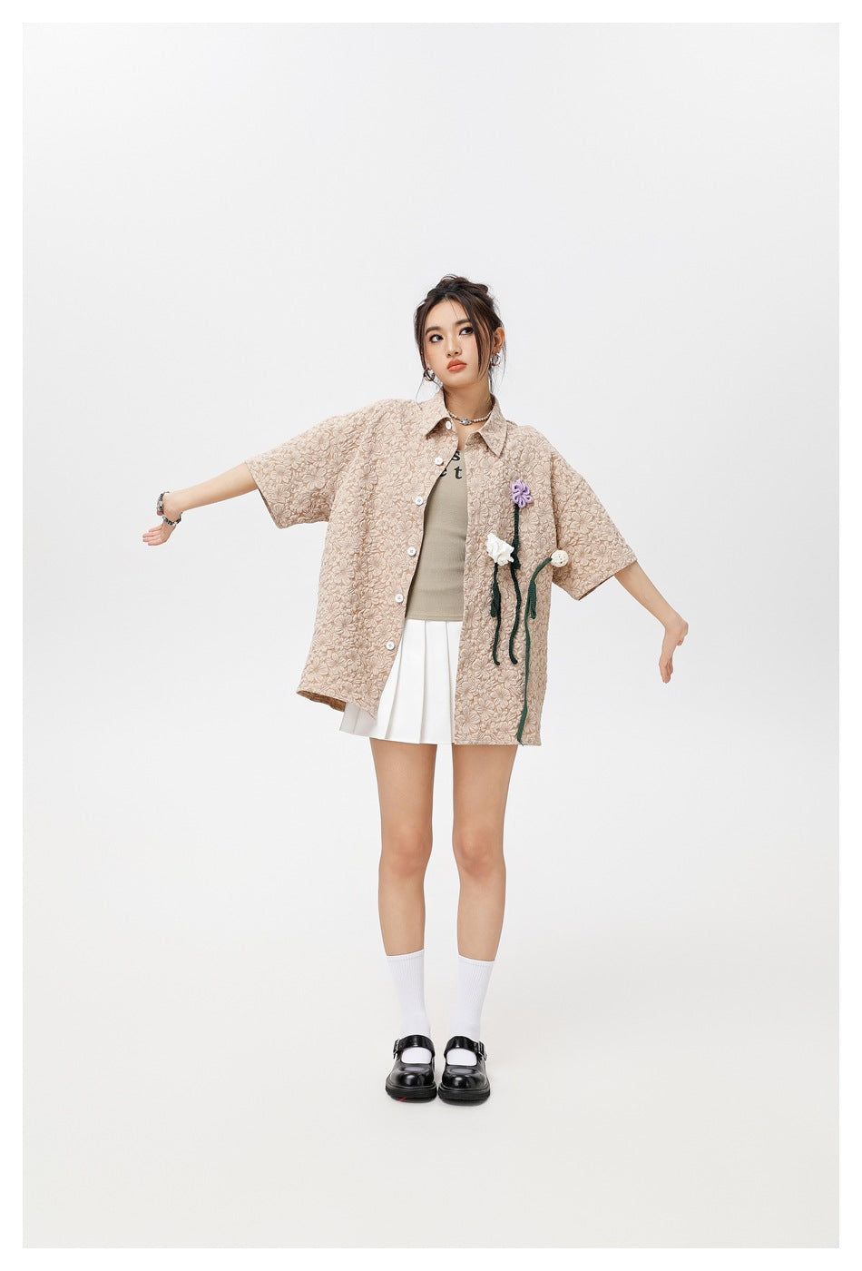 Full-length model wearing a loose beige floral-textured shirt with flower appliqués, a white pleated skirt and black shoes.