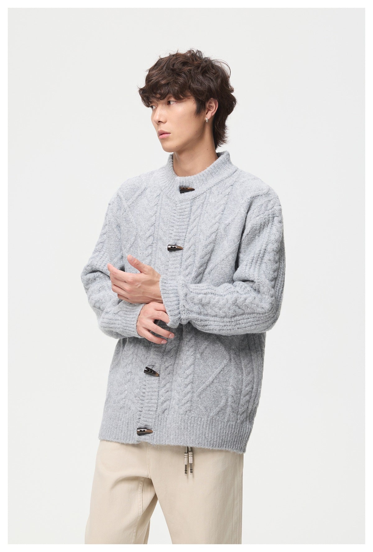 Three-quarter view of a model in a light gray cable-knit toggle cardigan and cream trousers.