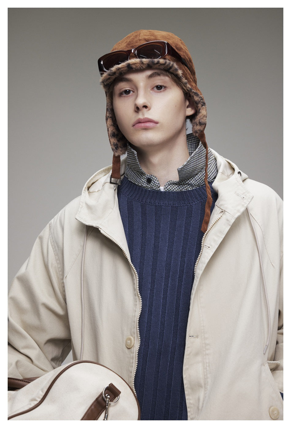 Close view of a model wearing an off-white hooded jacket, navy ribbed sweater, checked shirt, and brown fur-trimmed hat.