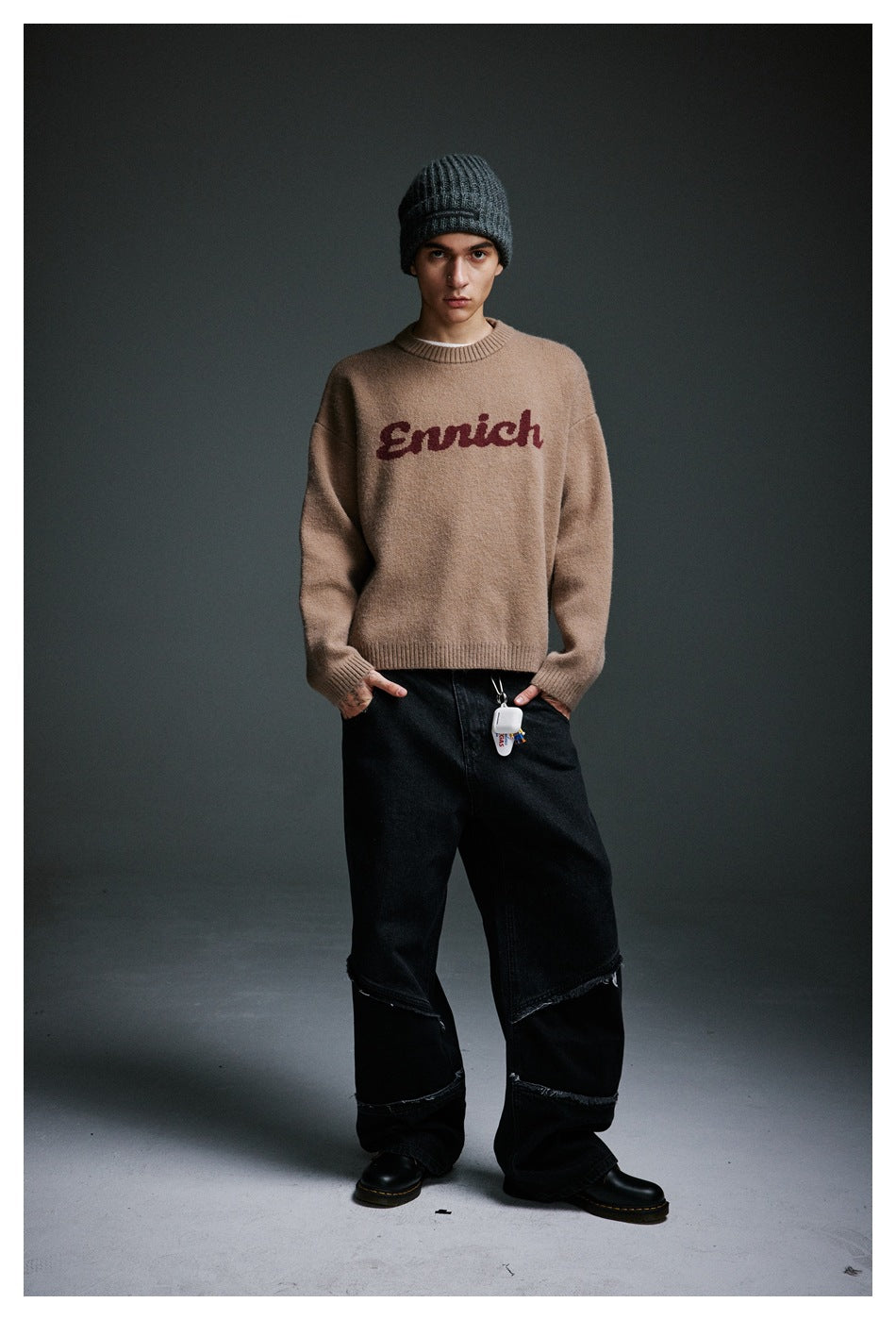 Full-length view of a model wearing a taupe sweater with burgundy script lettering, black jeans, and a dark beanie.