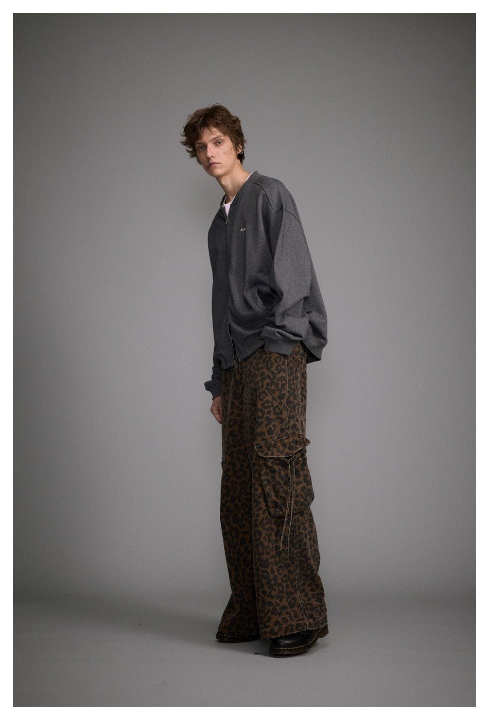 Model wearing an oversized charcoal gray zip-up jacket with a small chest logo and brown leopard-print cargo pants.