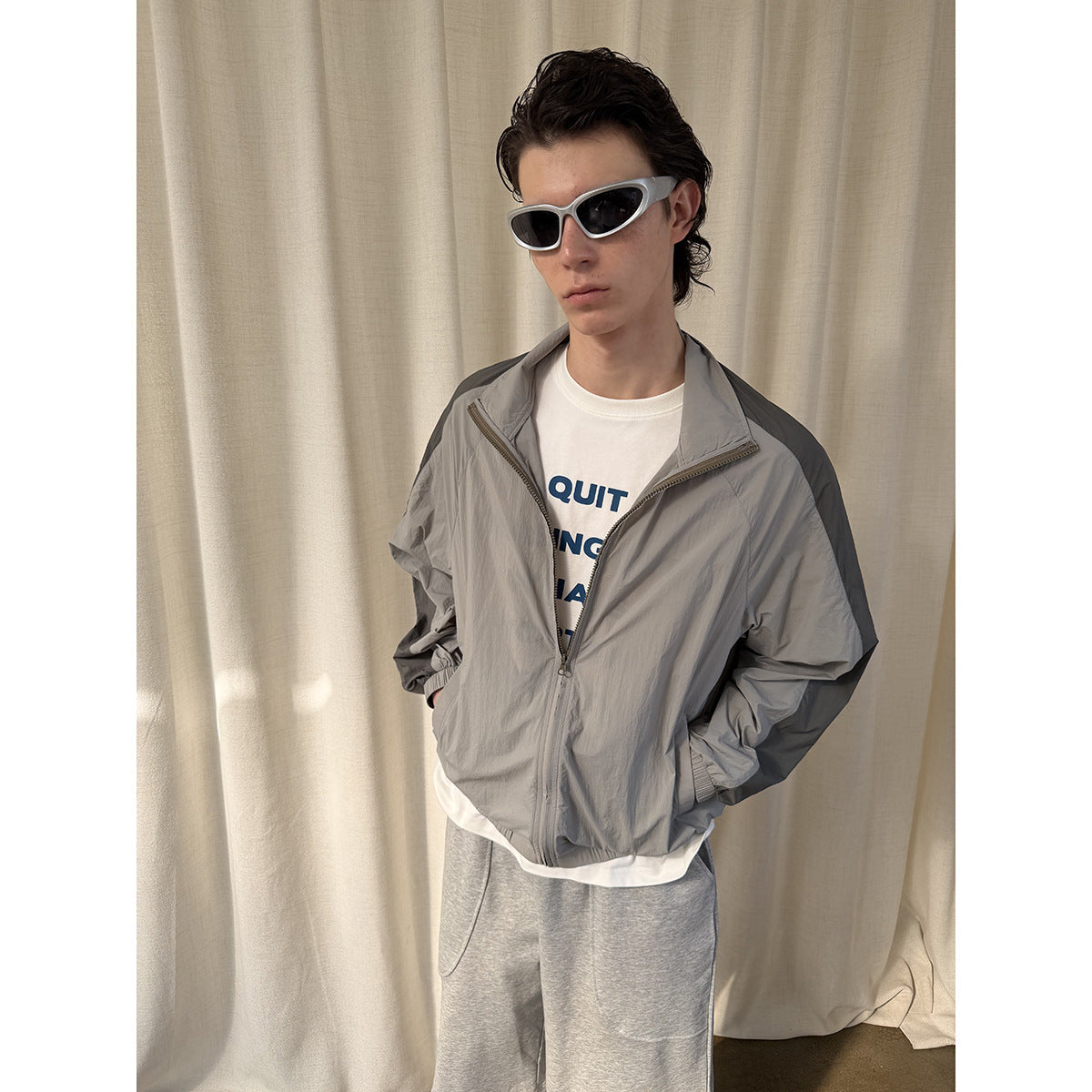 Model wearing a light gray jacket with dark sleeve panels, partly unzipped over a white graphic T-shirt.