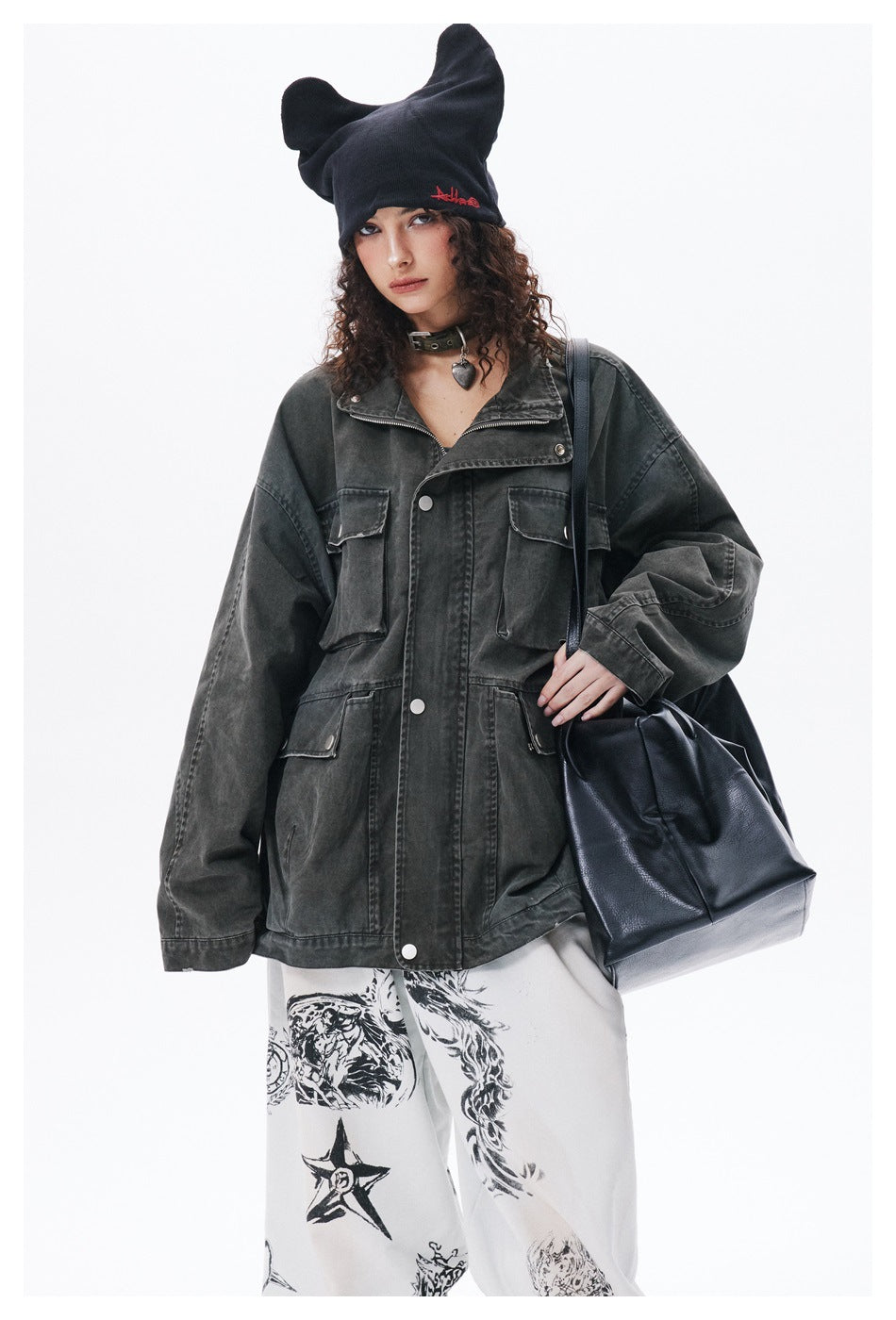 Model wearing a charcoal utility jacket and black ear-shaped beanie, with white graphic trousers partly visible.