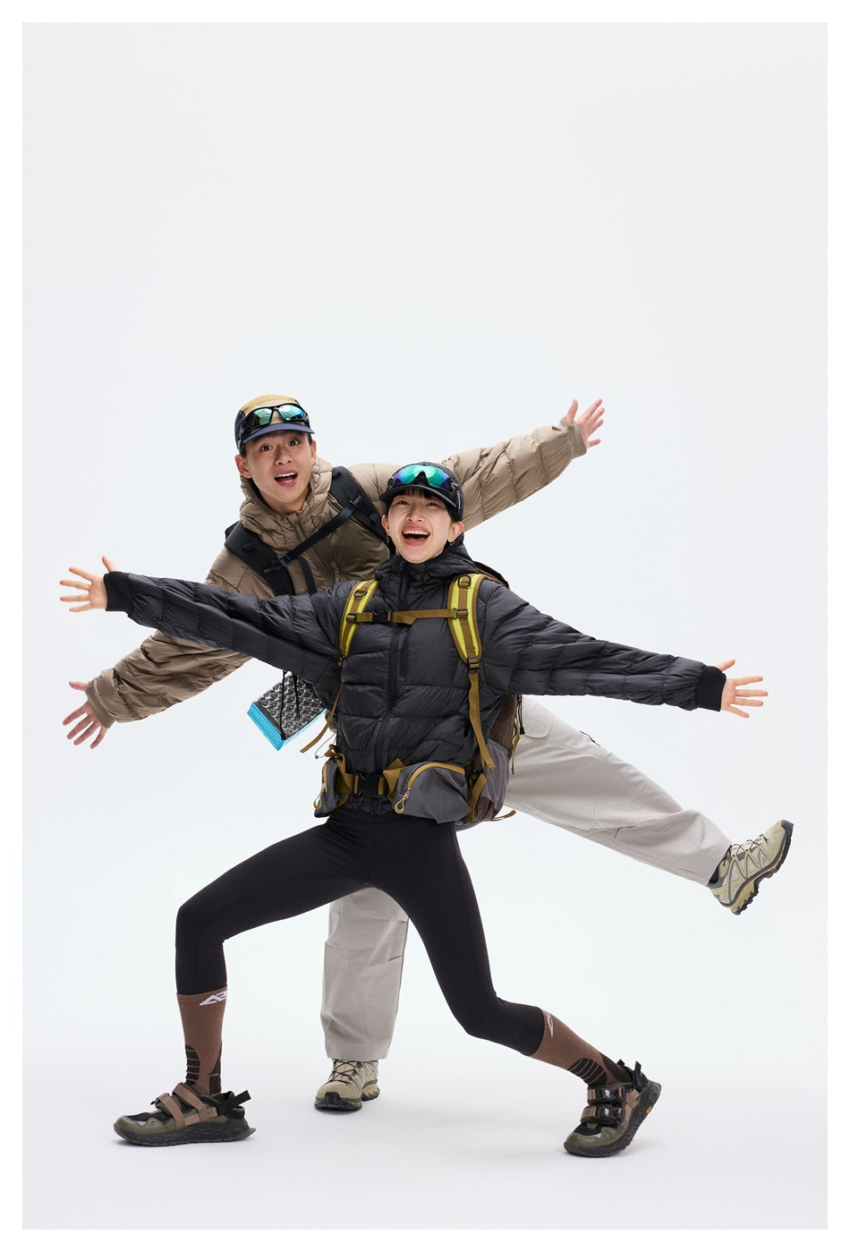 Two hikers pose in taupe and black hooded puffer jackets, backpacks on, with their arms spread against a white studio