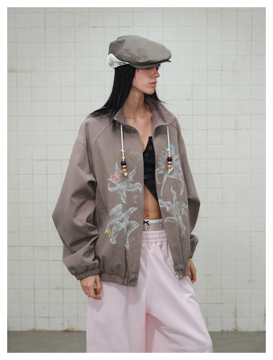 Model in side view wearing a taupe zip jacket with beaded collar drawcords and front print, with a cap and pink pants
