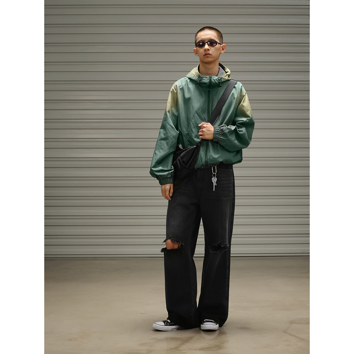 Man wearing a dark green hooded zip jacket with elastic cuffs, black ripped jeans and a crossbody bag.