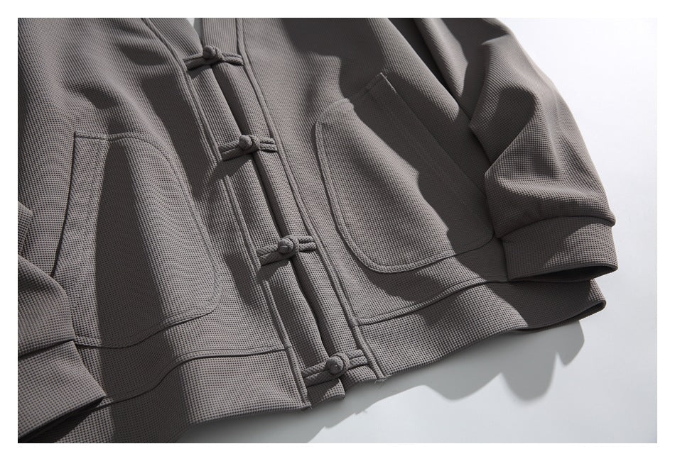 Close-up of a gray textured jacket front showing knotted frog closures, patch pockets, and a short sleeve.
