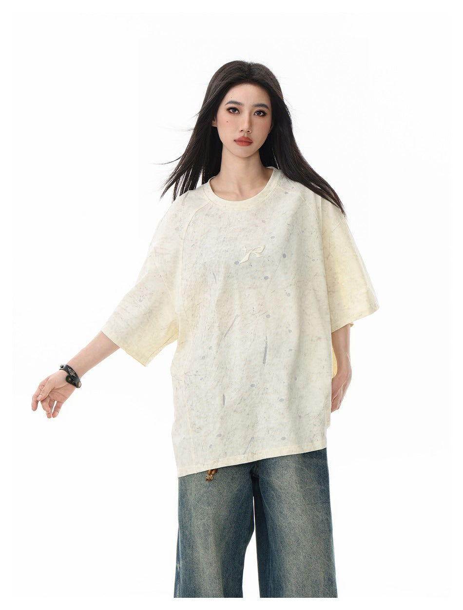 Model in oversized beige short-sleeve T-shirt with dropped shoulders and splatter marks, with wide-leg faded jeans
