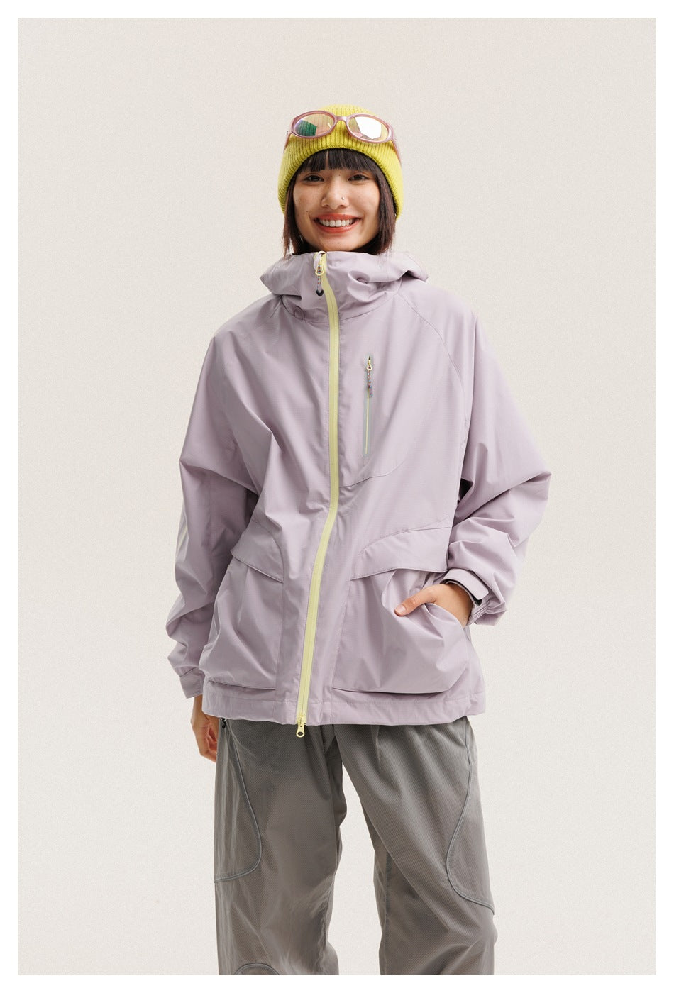 Front view of a woman wearing a pale lilac hooded jacket with a lime zipper and gray pants.