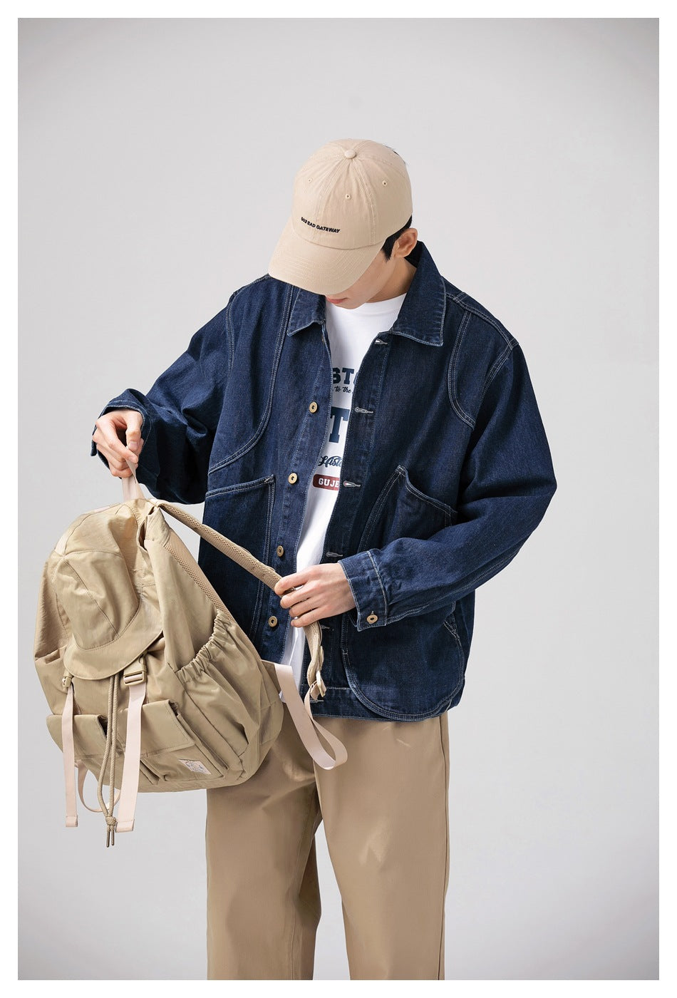 Closer view of a model wearing a dark indigo denim jacket and holding a beige backpack.