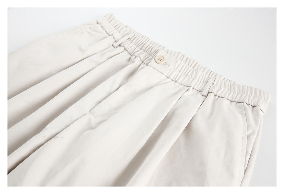 Off-white trousers laid flat, showing the gathered elastic waistband, button, belt loops, pleats, and front pockets.