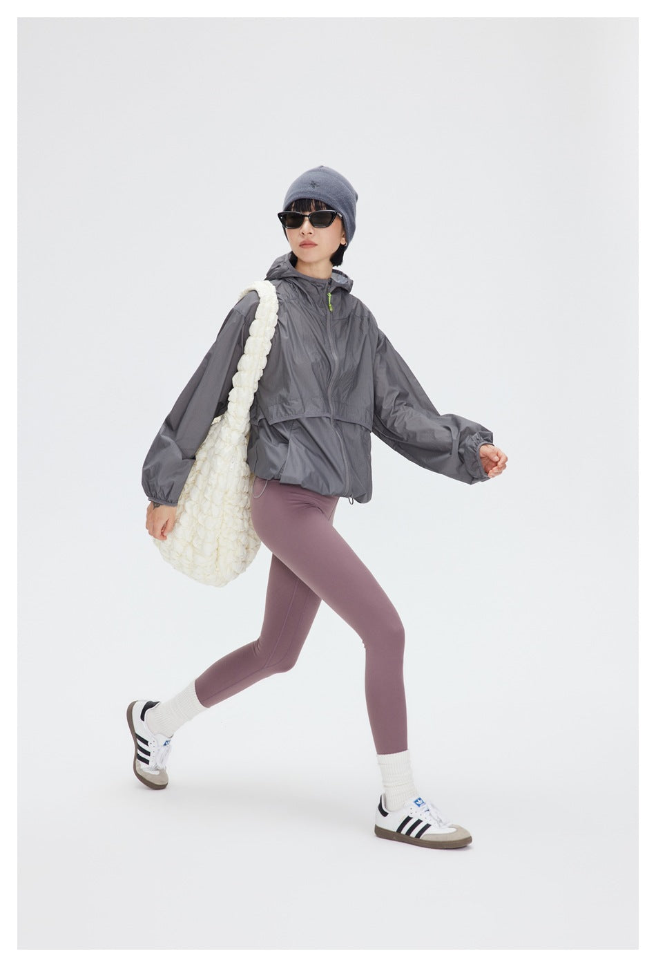 Model wearing a gray zip-front hooded windbreaker and mauve leggings, walking with a textured white shoulder bag.