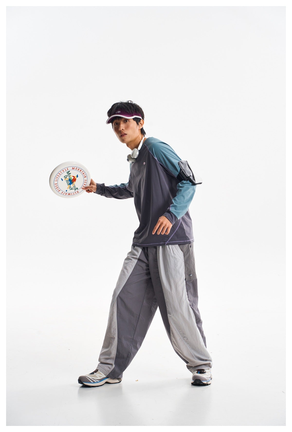 Model in a charcoal shirt with teal sleeves and two-tone gray pants, holding a flying disc.