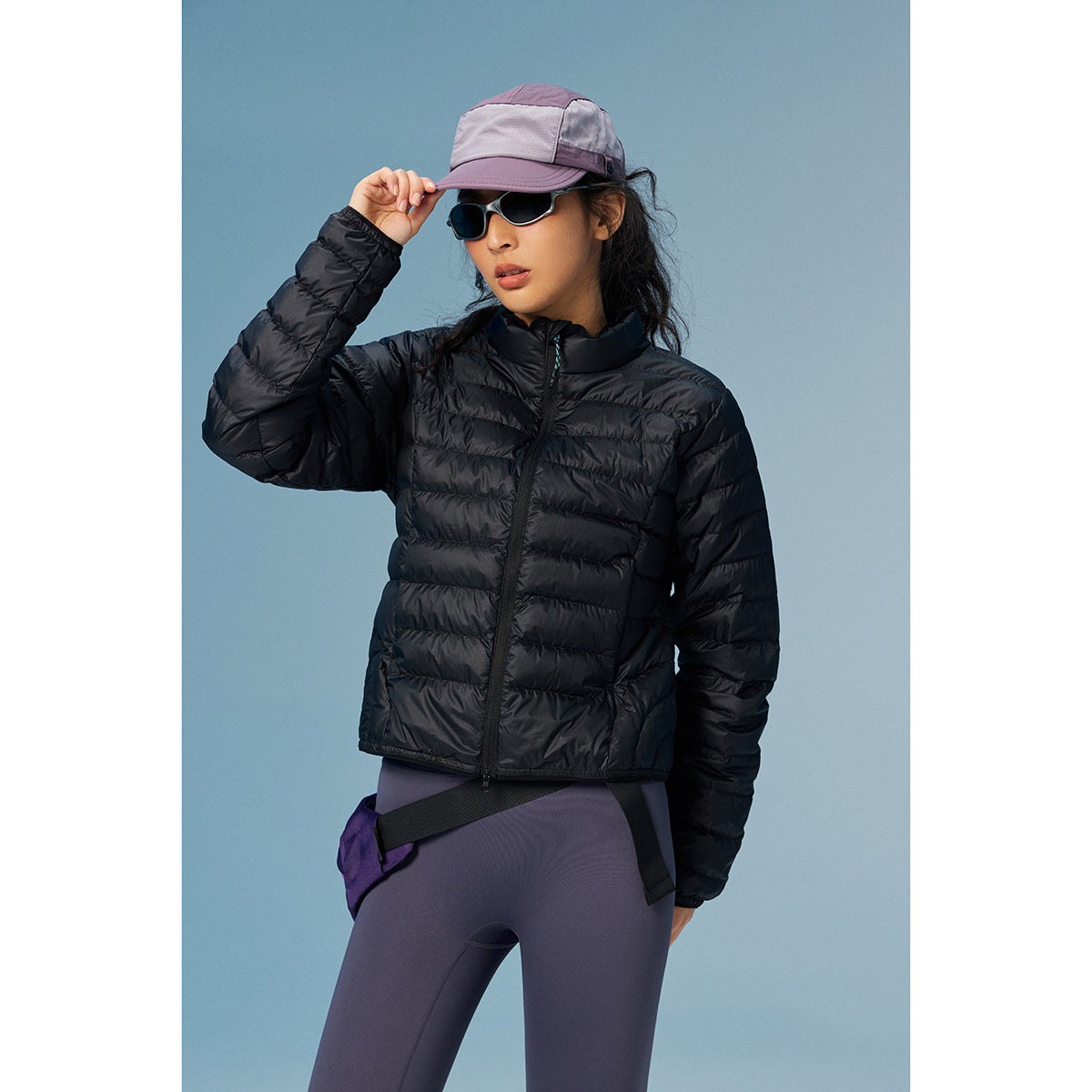 Model wearing a black quilted zip jacket with a high collar, purple leggings, and a lavender cap.