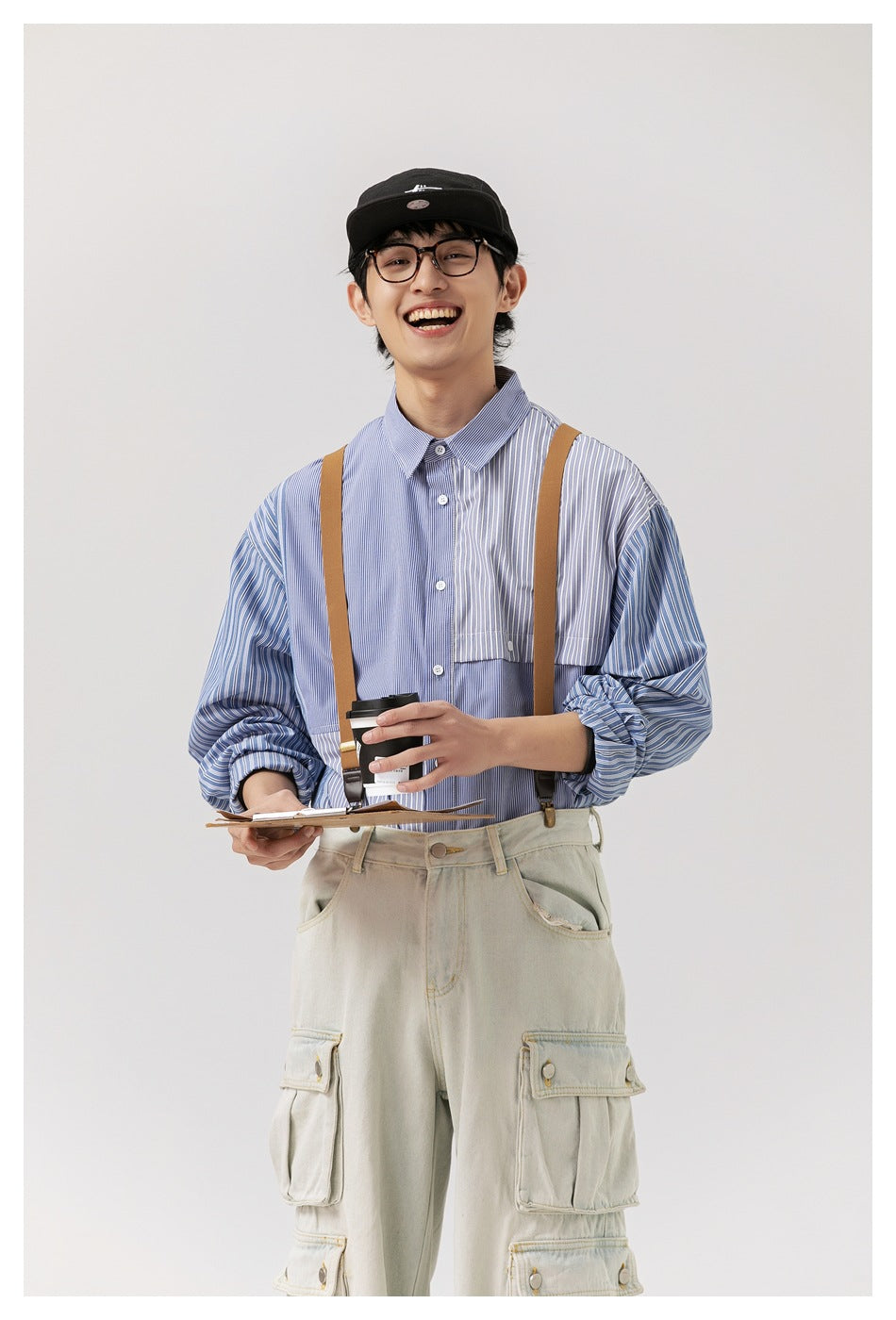 Model smiling in a blue patchwork striped button-up shirt with brown suspenders and pale cargo pants.