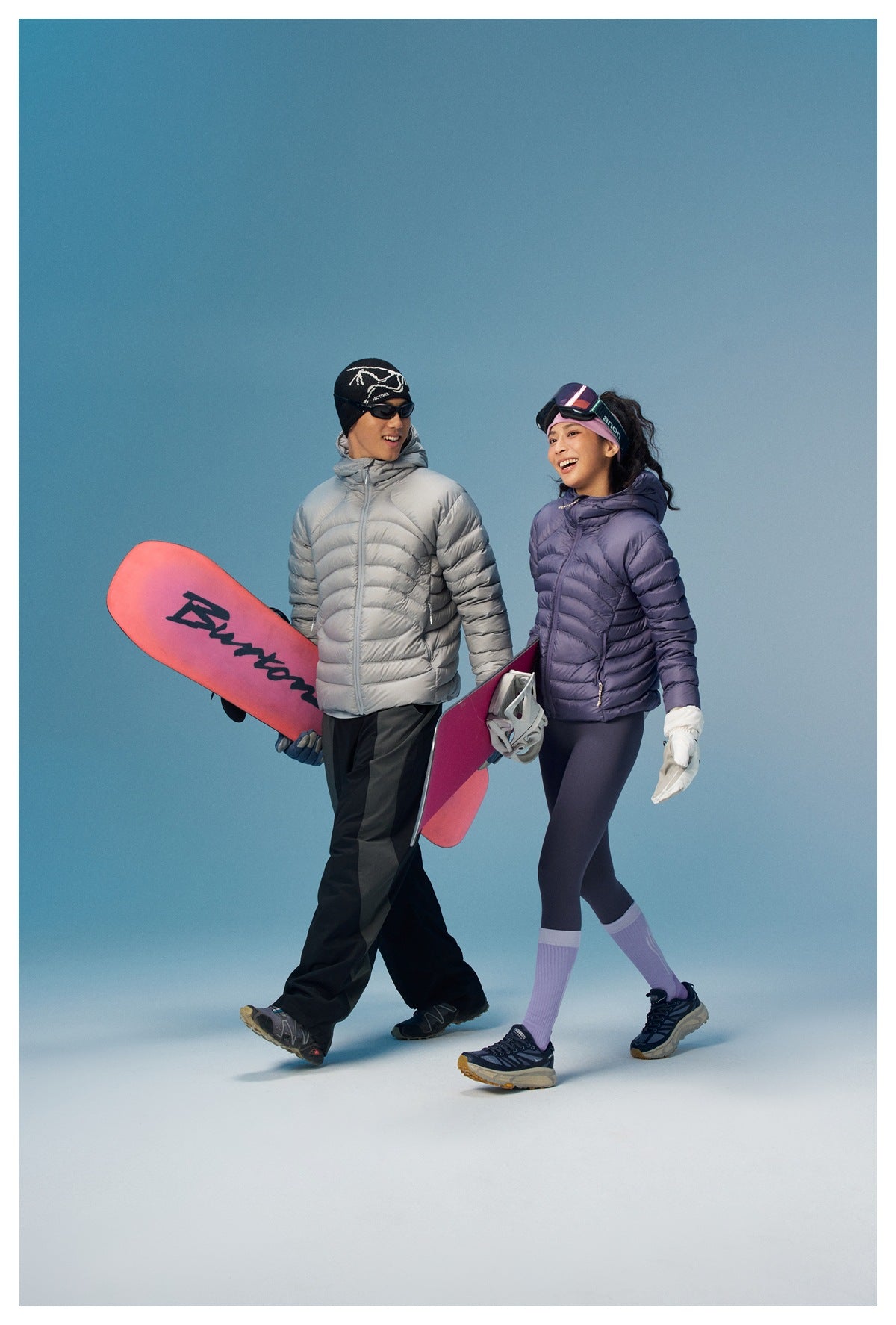 Two people walk with snowboards, wearing light gray and lavender quilted puffer jackets.