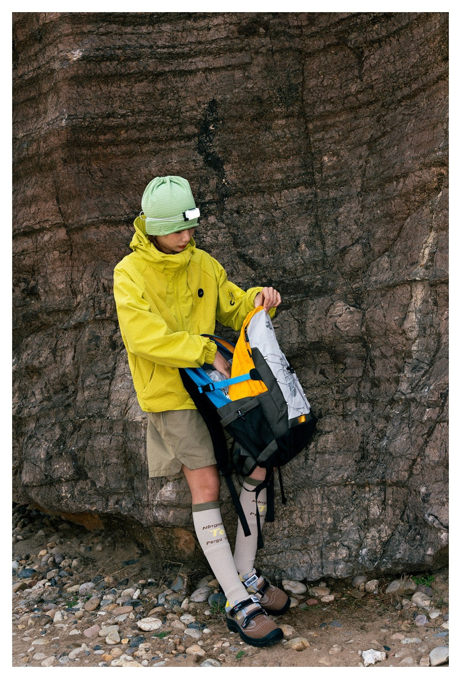 Person in a bright yellow hooded jacket, khaki shorts and green beanie, handling a multicolor backpack by a rocky wall.