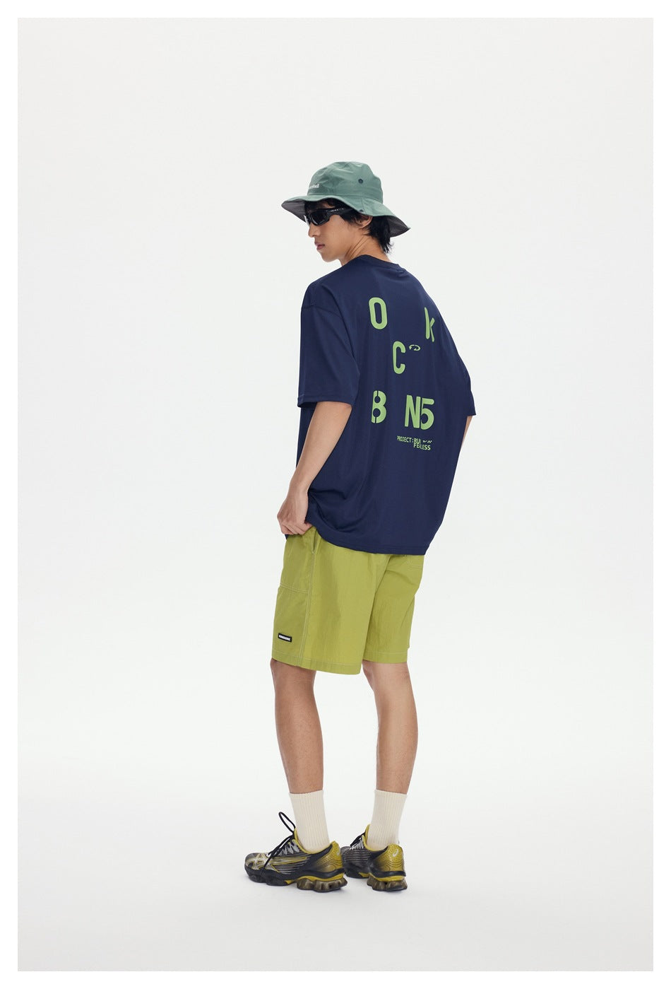 Model wearing an oversized navy graphic T-shirt, lime shorts, and a sage bucket hat, shown from the back.