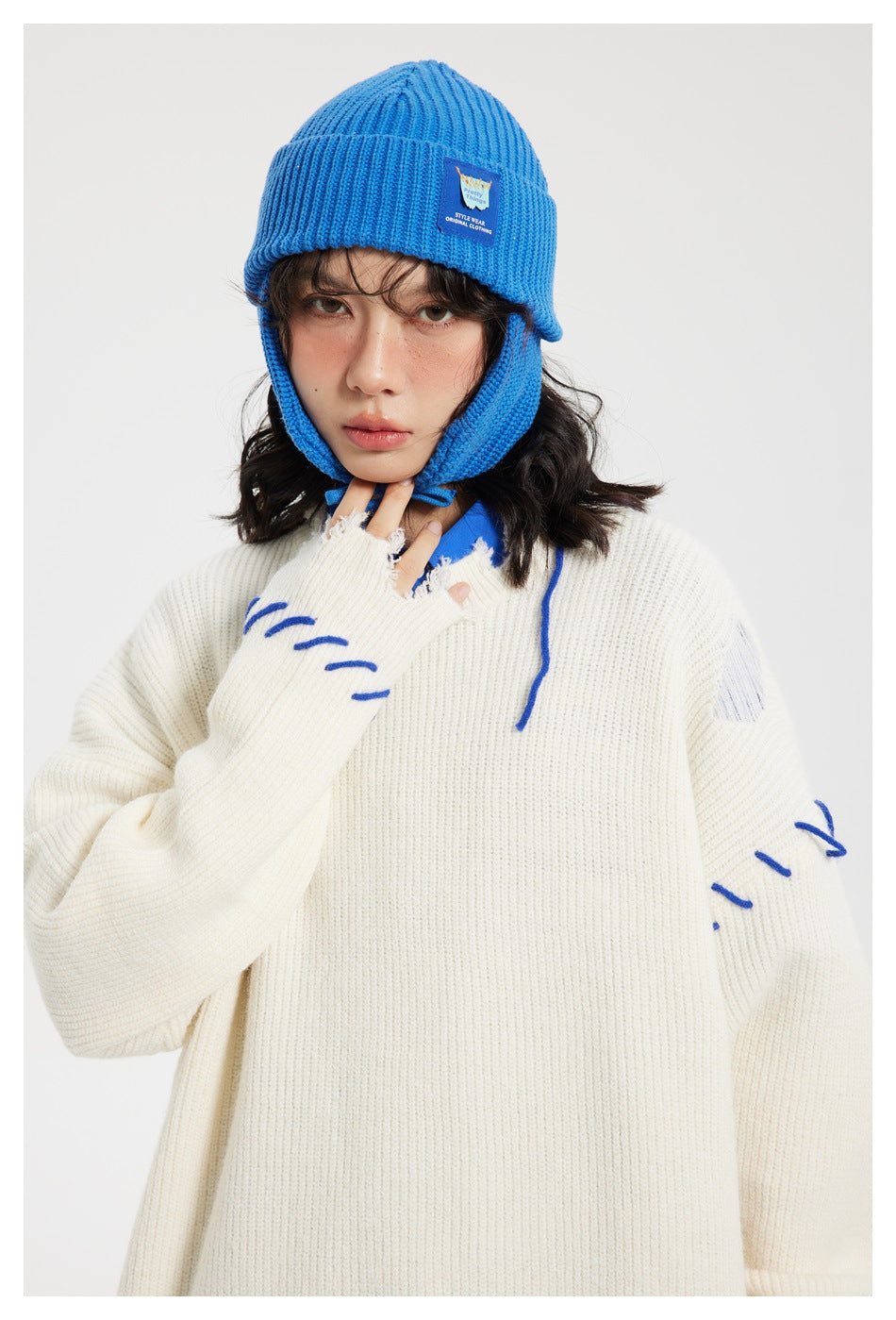 Close-up of a model wearing an ivory sweater with blue stitch accents and a bright blue ribbed earflap beanie.
