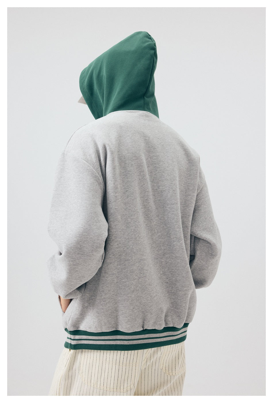 Back view of a person wearing a gray hoodie with a dark green hood and striped green hem, paired with pinstriped pants.