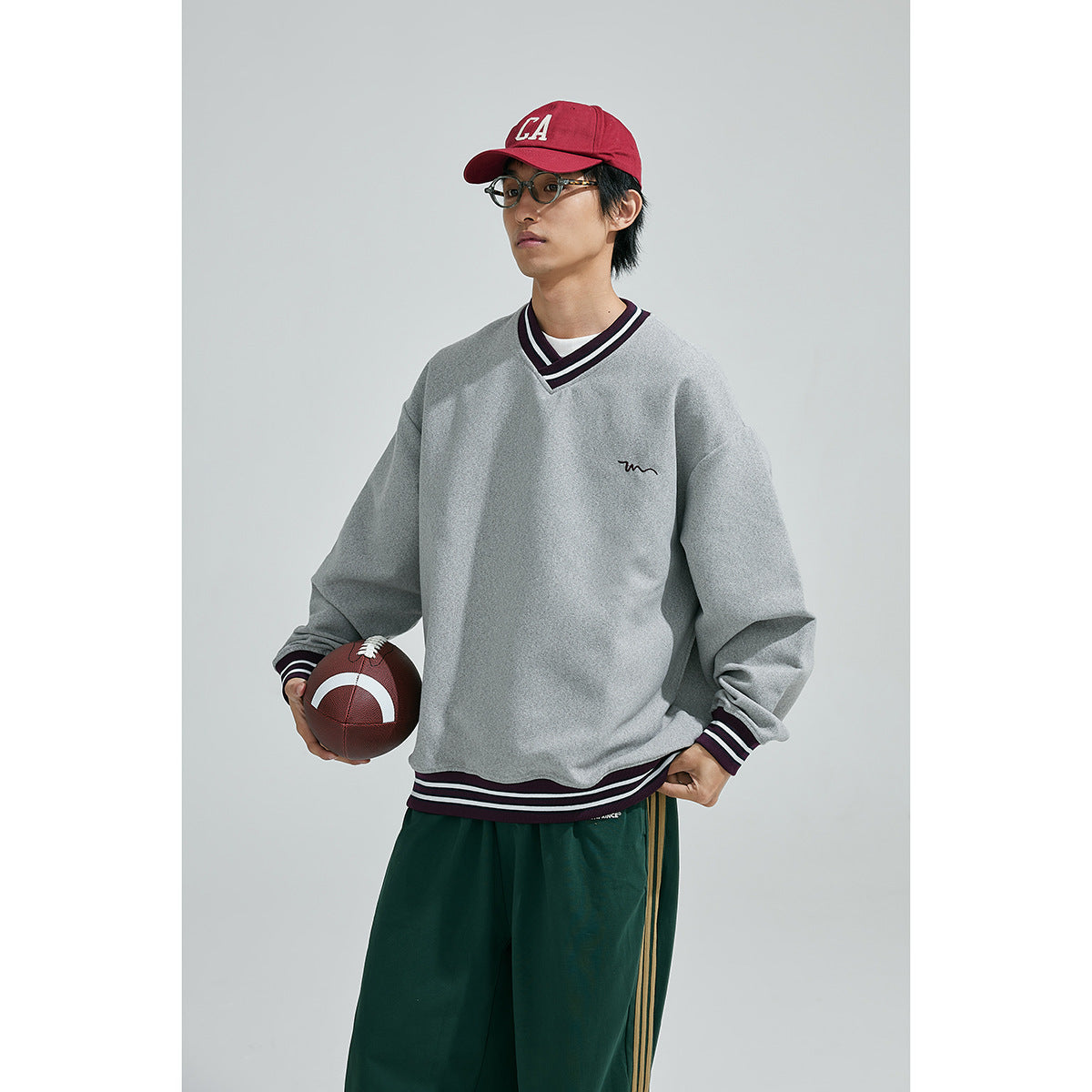 Three-quarter view of a model in a gray striped-trim V-neck sweatshirt and green track pants, holding a football.