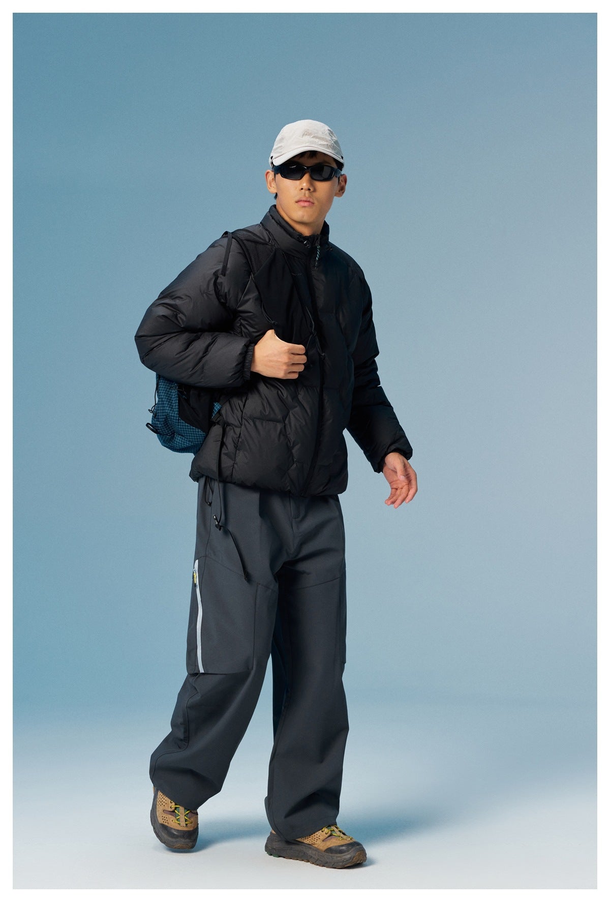 Model walking in a black quilted puffer jacket with stand collar, gray pants, cap, sunglasses and a backpack