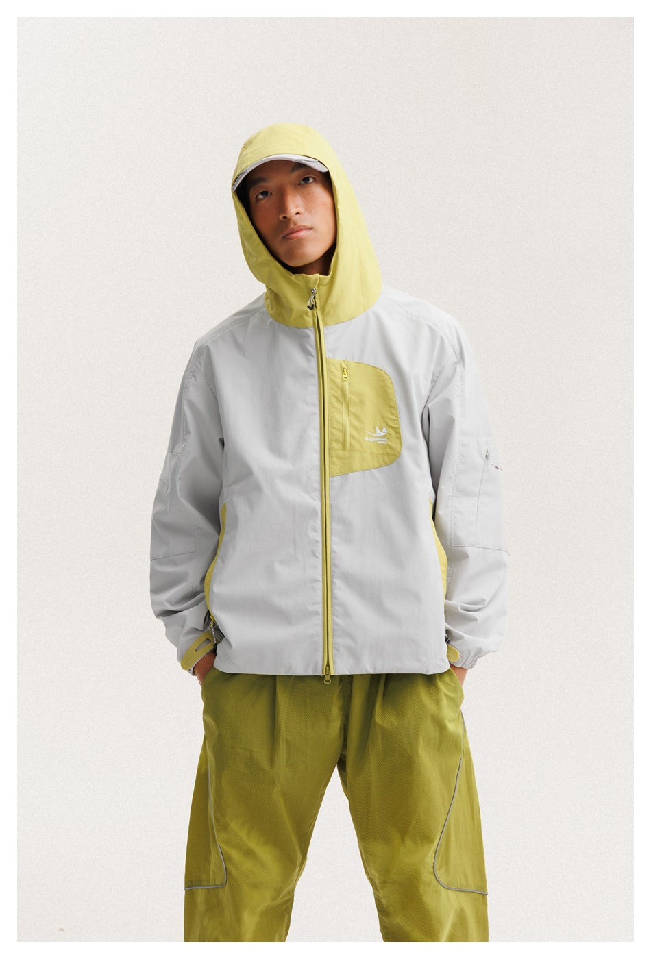 Front view of a model wearing a light gray jacket with a chartreuse hood, chest pocket and trim, plus chartreuse pants.