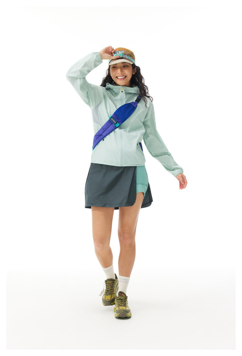 Model wearing a pale mint hooded jacket and dark teal skirt, with a blue crossbody bag and hiking shoes.