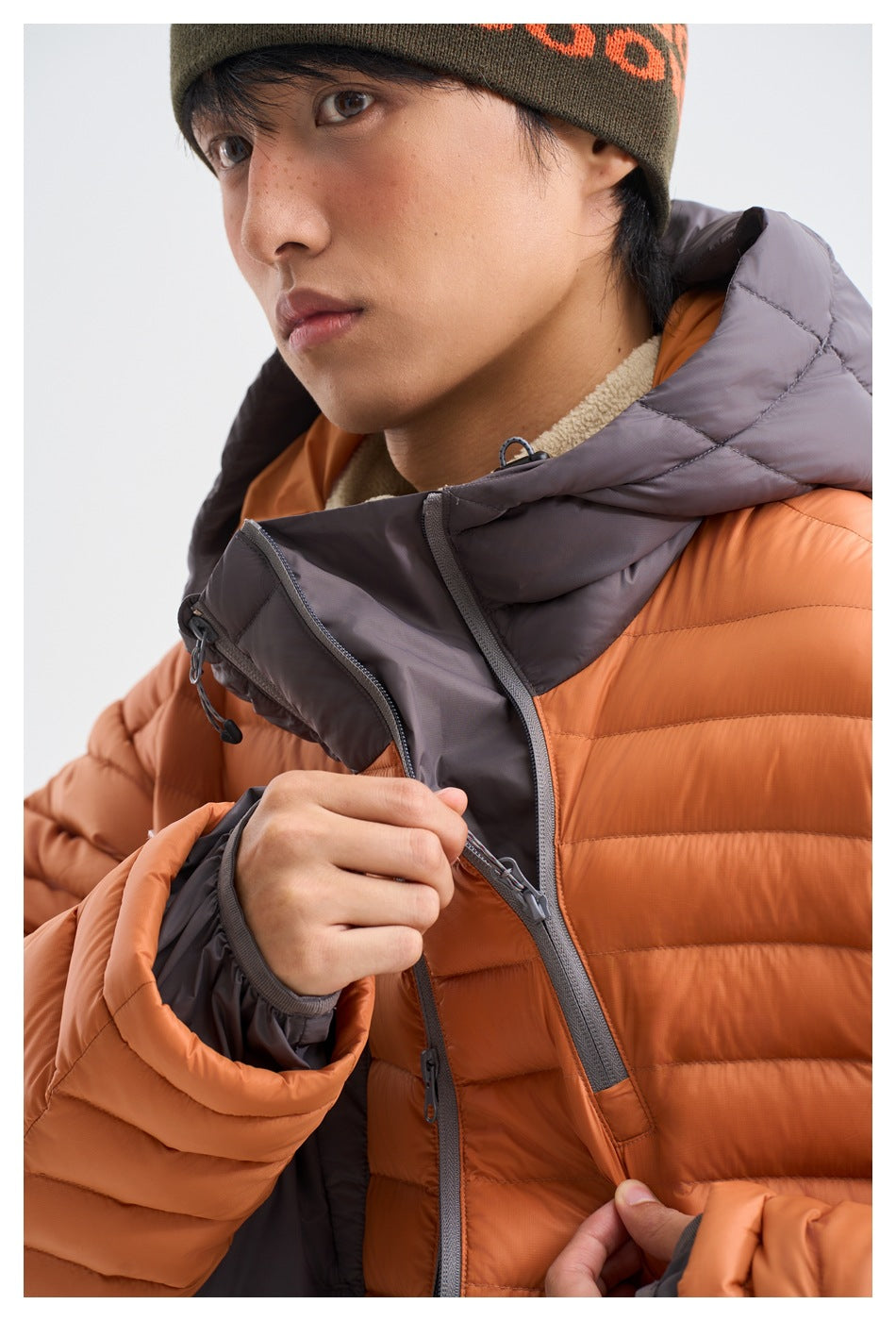 Close-up of a model opening the gray-lined hood and front zipper of an orange puffer jacket.