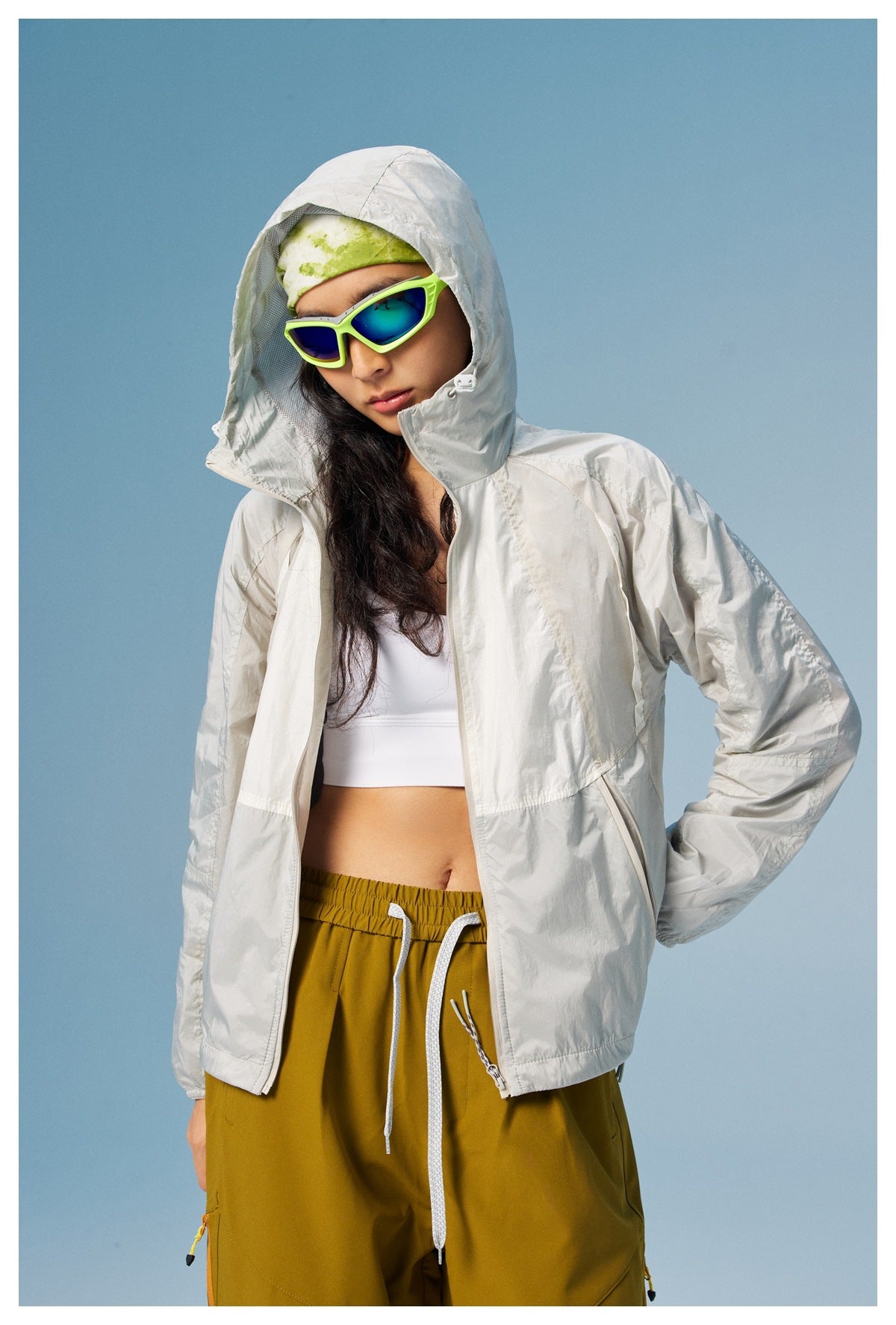 Woman wearing a light gray hooded jacket over mustard pants, with the hood up and jacket partly open.