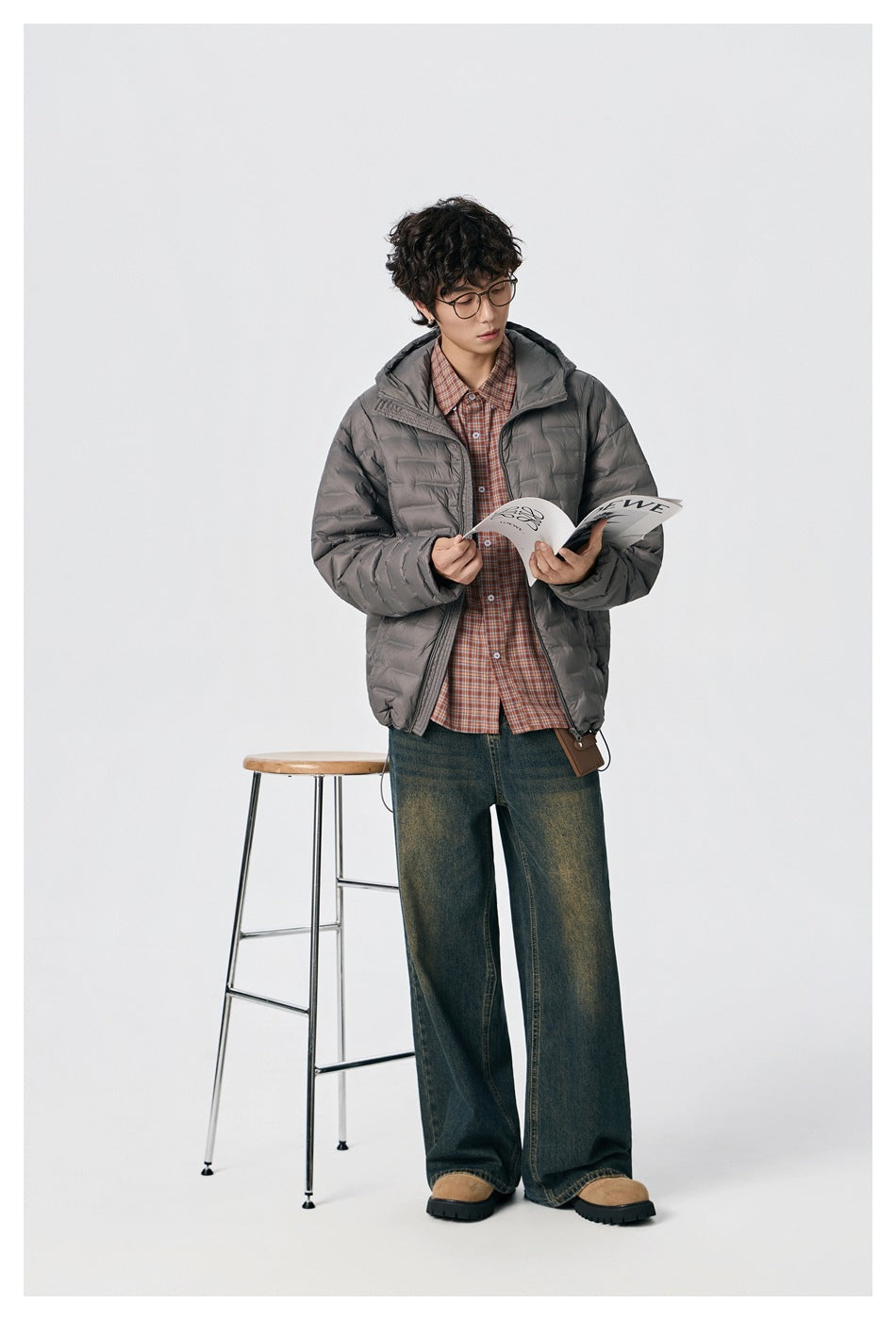 Model wearing an open gray quilted hooded jacket over a plaid shirt, holding an open booklet.