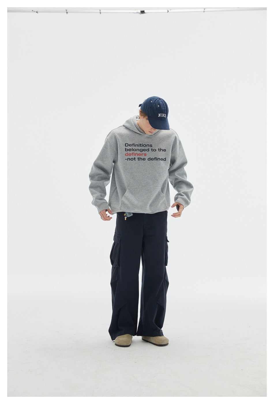 Model looks down while wearing a gray slogan hoodie, navy cargo pants, and blue cap, shown full-length from the front.