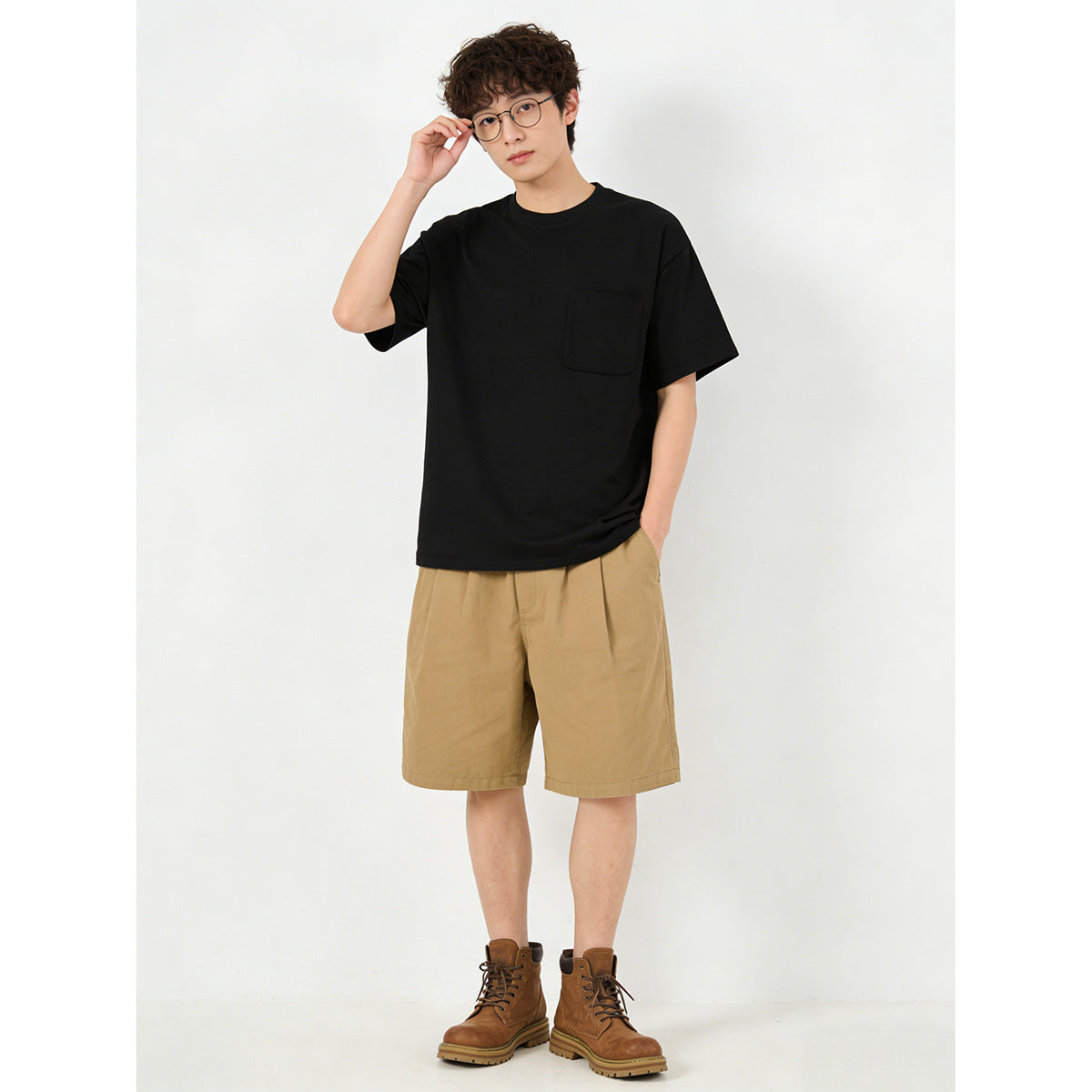Model wearing a loose black crew-neck T-shirt with a chest pocket and tan shorts.