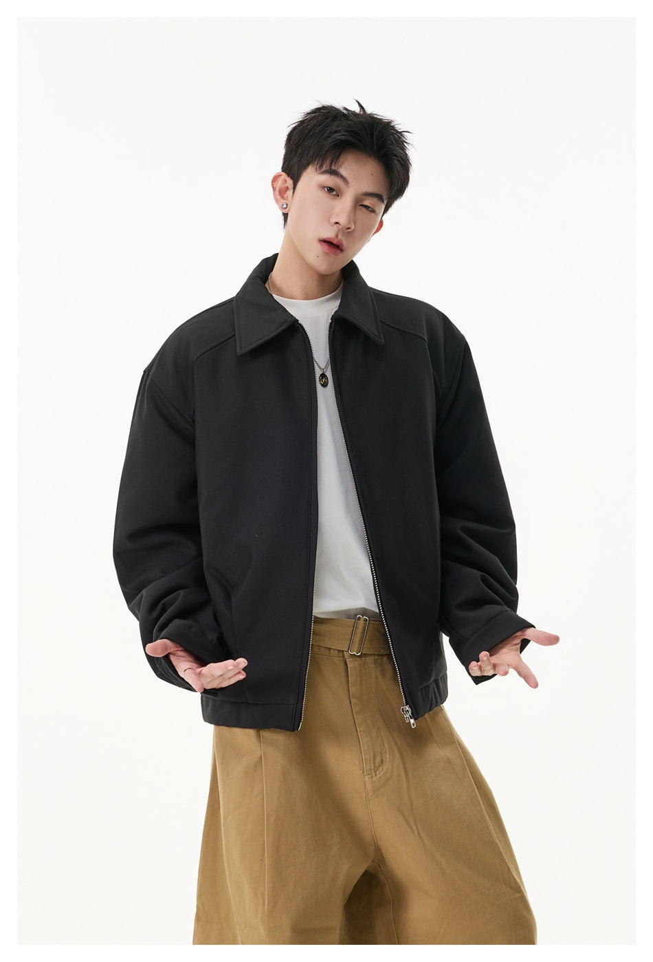 Model wearing an open black collared zip jacket over a white shirt and tan trousers.