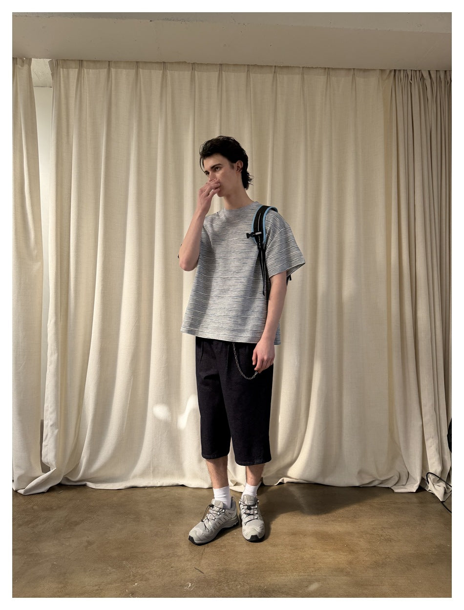 Model with hand near face wearing a gray striped short-sleeve T-shirt with dark shorts, in front of a cream curtain