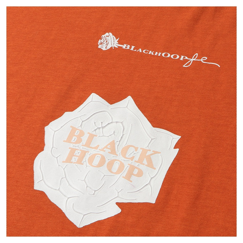 Close-up of an orange garment with a raised white rose graphic and pale lettering reading “BLACK HOOP.”