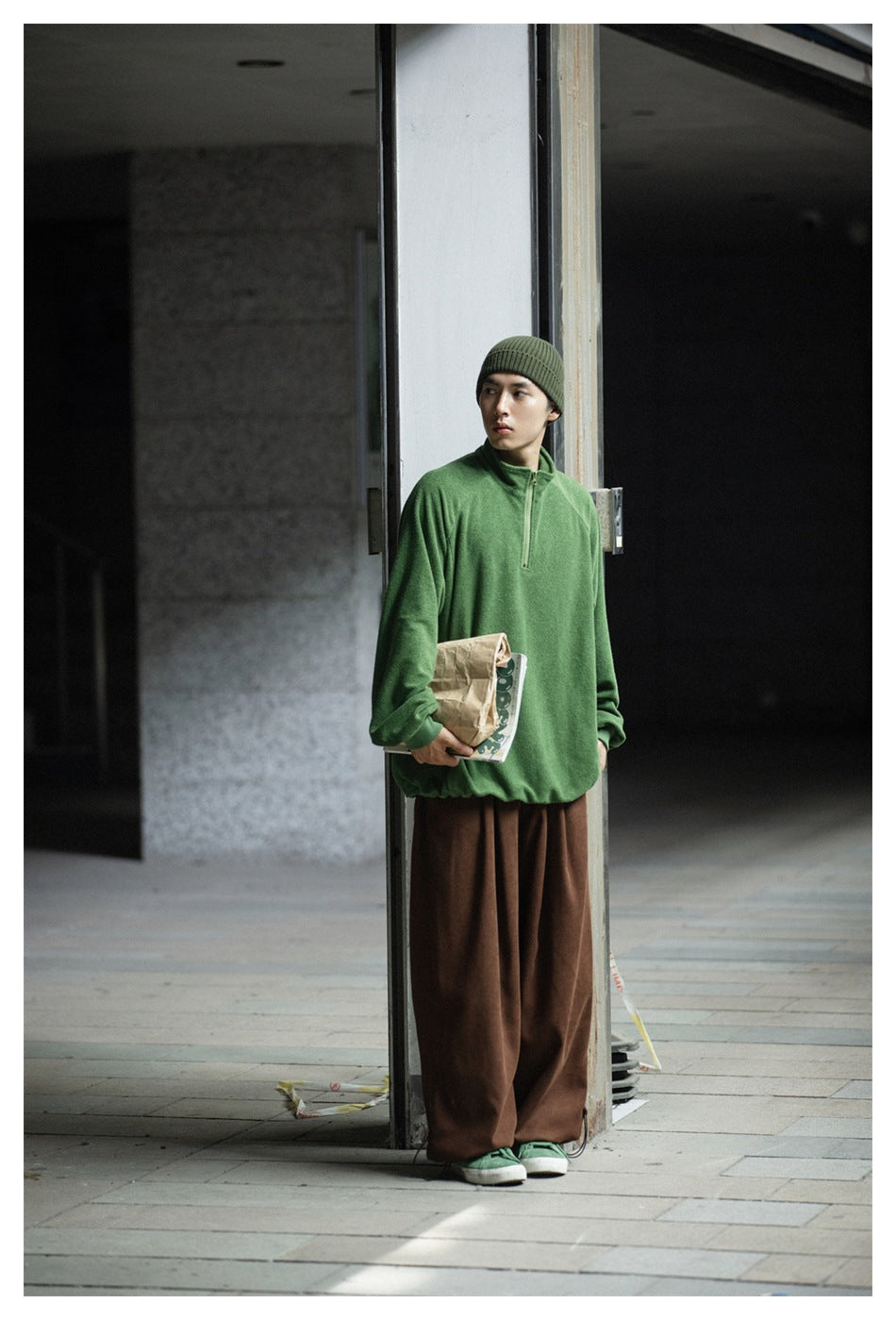 Model standing by a pillar in brown wide-leg pants, a green fleece-like half-zip pullover and a green beanie