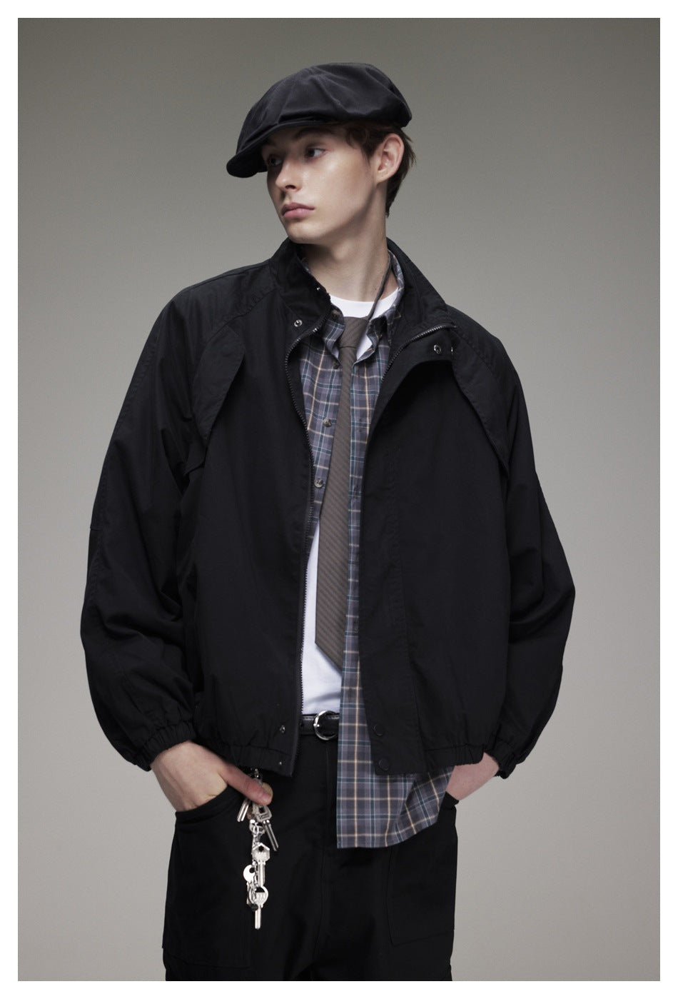 Model wearing an open black jacket over a plaid shirt, white tee and tie, with a black cap.
