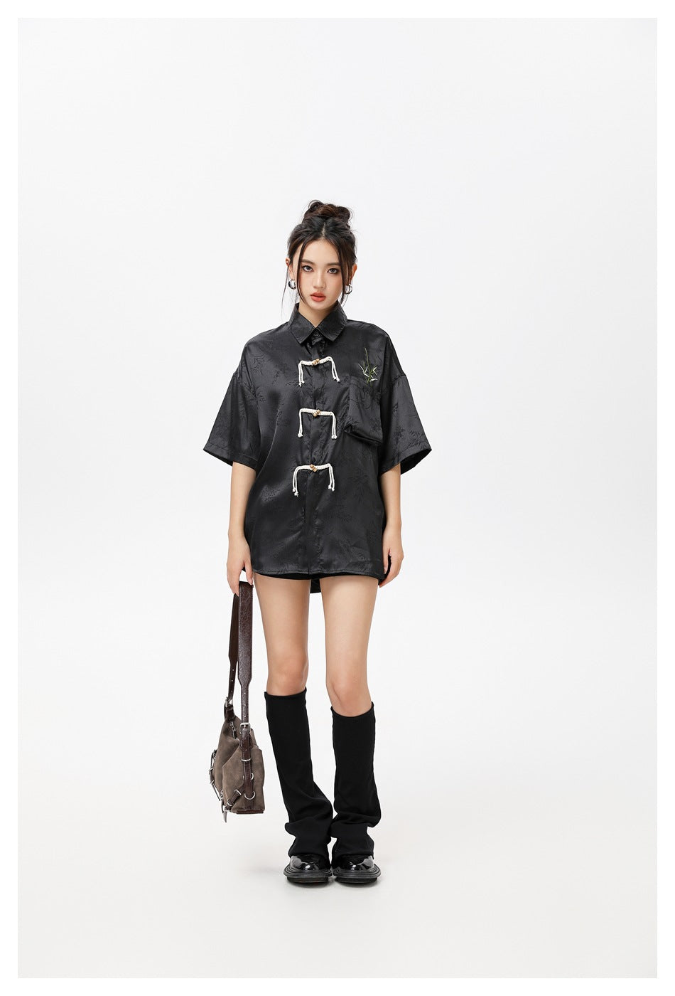 Model wearing an oversized black short-sleeve shirt with three cream rope frog closures, a chest pocket, and bamboo
