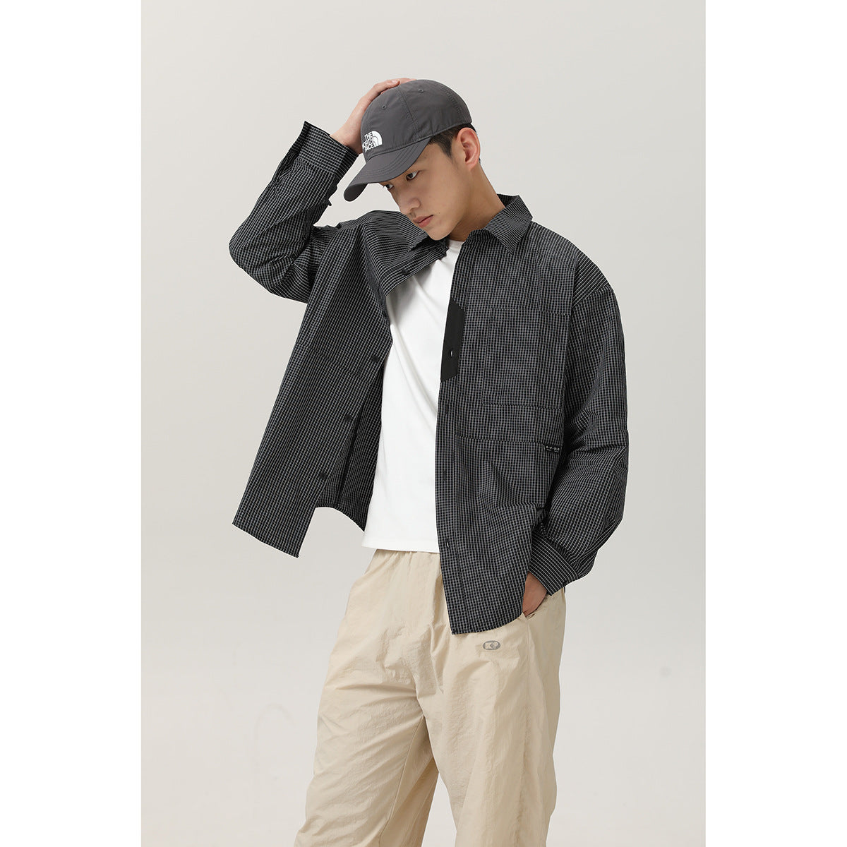 Model wearing an open charcoal micro-check shirt over a white tee, shown with loose beige trousers.