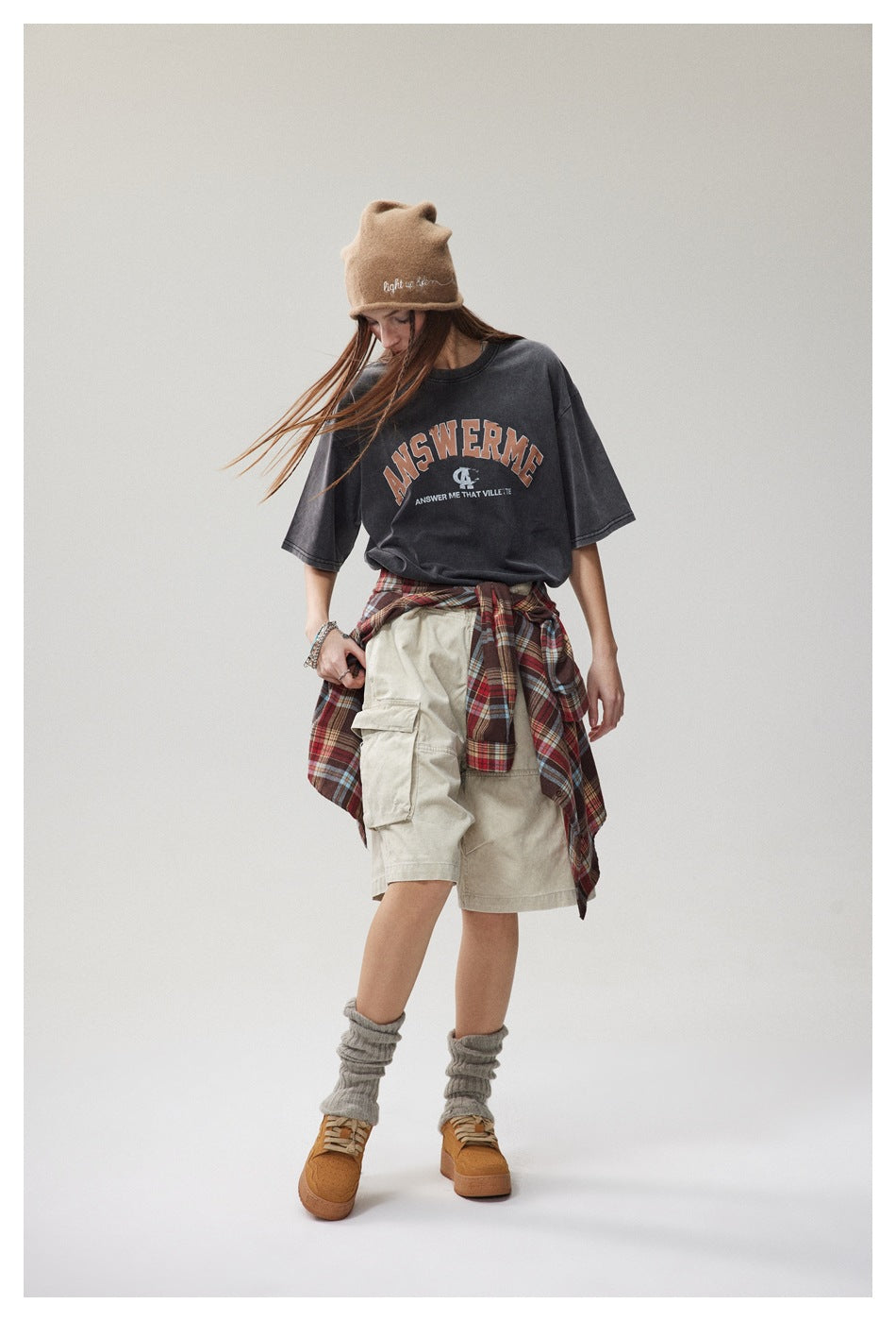 Model looking down in washed black ANSWERME T-shirt with beige cargo shorts, plaid shirt at waist and beanie