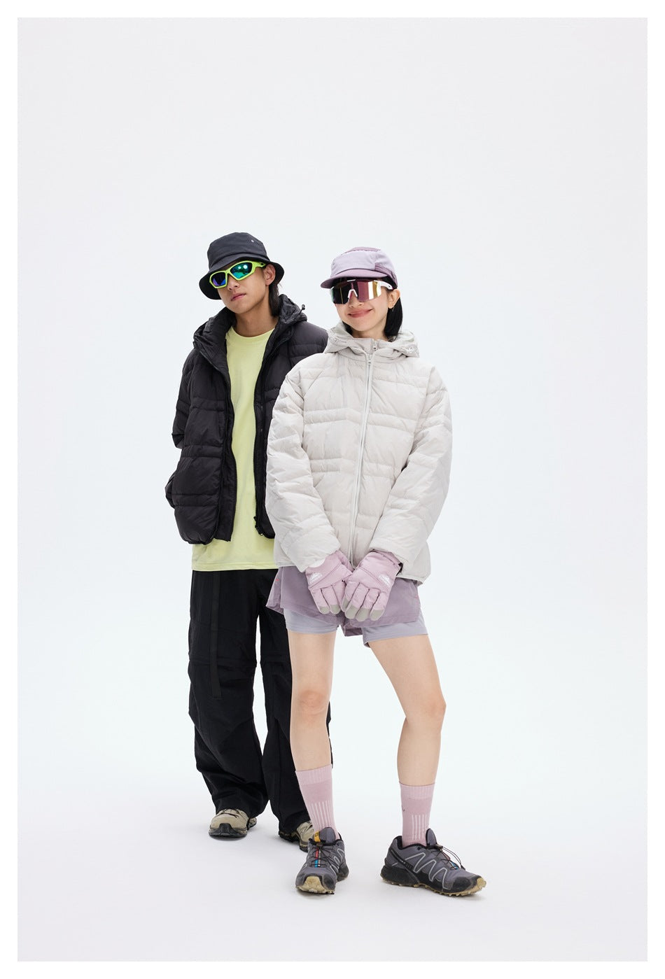 Two models pose together in black and light gray quilted hooded jackets, with hats and mirrored sunglasses.