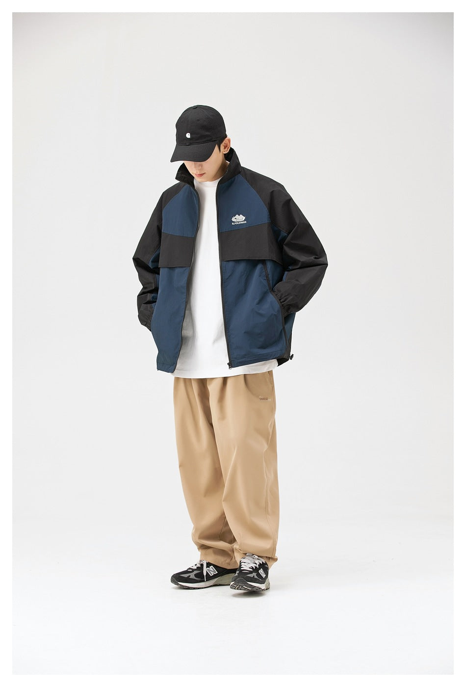 Model wearing navy and black color-block jacket open over a white tee with khaki trousers, looking down