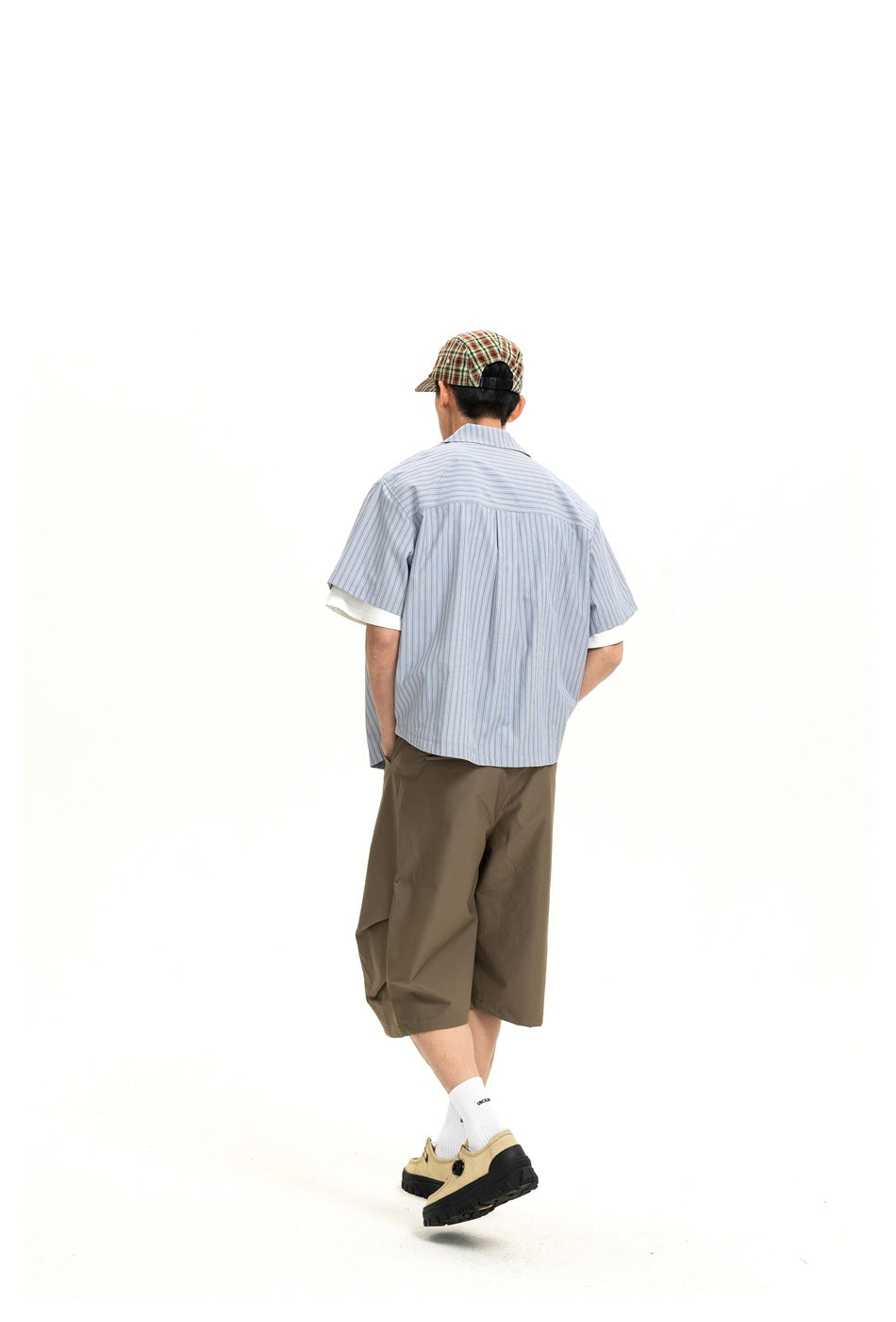 Back view of a man wearing a blue striped short-sleeve shirt with back yoke, olive shorts and a plaid cap