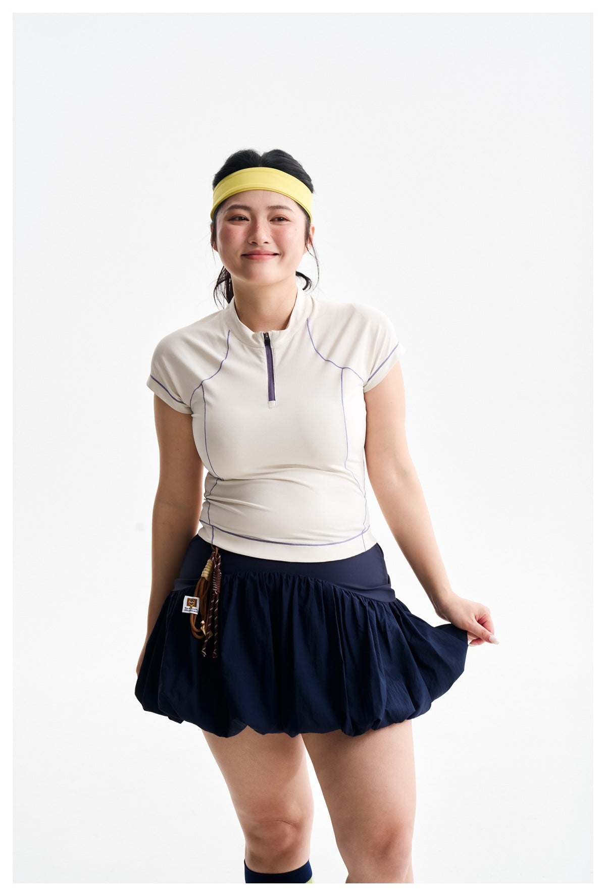 Model wearing an ivory zip-neck top with purple seam accents and a navy bubble skirt.