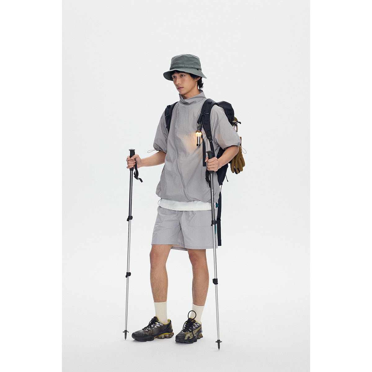 Model wearing a light gray short-sleeve zip jacket and matching shorts, shown with hiking gear and poles.